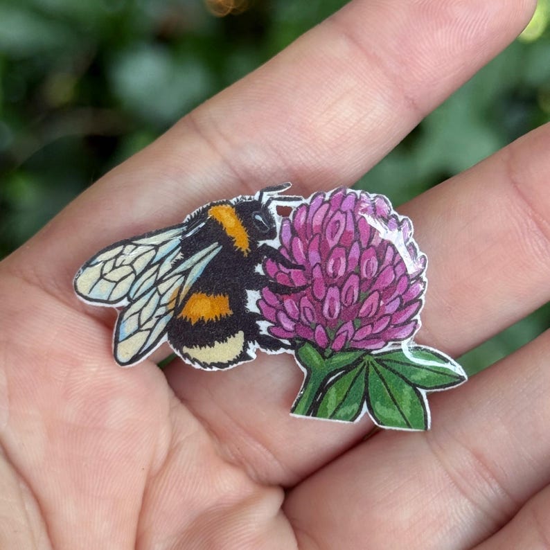 Puede incluir: Un pin plano que presenta una ilustraci&oacute;n detallada de un abejorro peludo con rayas negras y amarillas y alas transl&uacute;cidas, colocado junto a una flor de tr&eacute;bol de color p&uacute;rpura vibrante con hojas verdes, con un acabado brillante y claro.