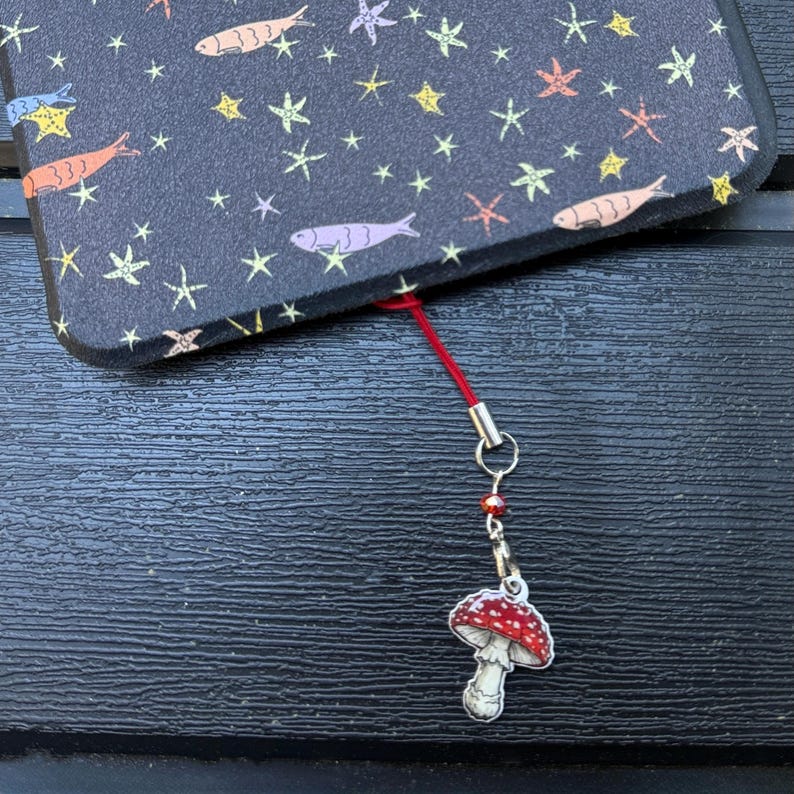 May include: A black phone case with a pattern of fish and stars, connected to a red lanyard with a mushroom charm. The mushroom charm has a red cap with white spots and a white stem.