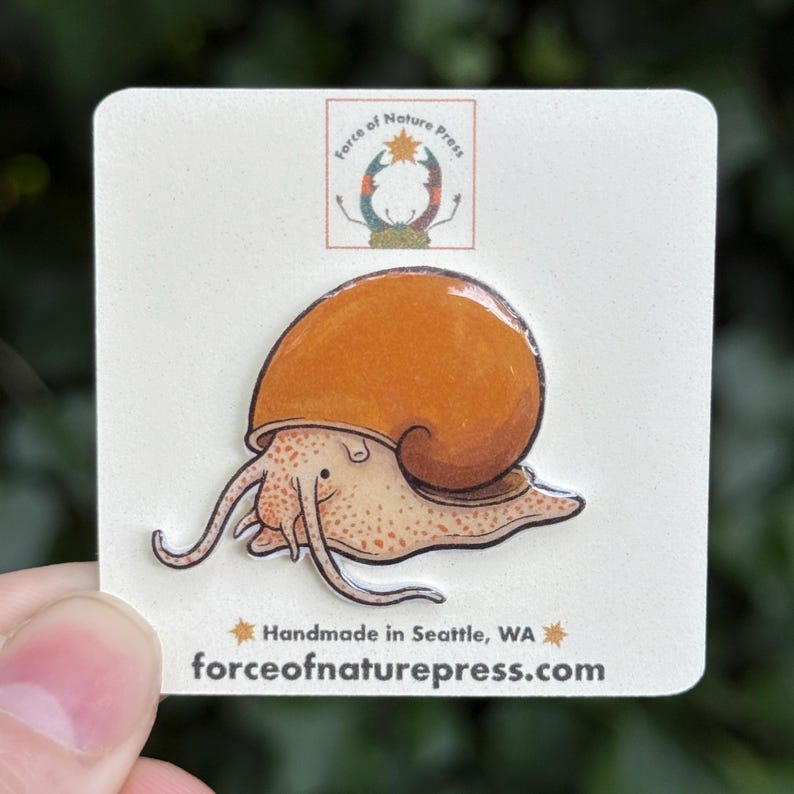 Puede incluir: Un pin de esmalte que muestra un caracol de color canela con una concha naranja grande y brillante. El pin est&aacute; sujeto a una tarjeta blanca con el texto Force of Nature Press, Handmade in Seattle, WA y forceofnaturepress.com impreso.