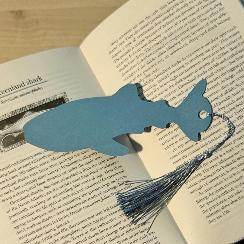 May include: A light blue shark-shaped bookmark with a silver tassel, resting on a book page. The bookmark is made of paper and has a hole for the tassel. The book is open to a page with text about Greenland sharks.