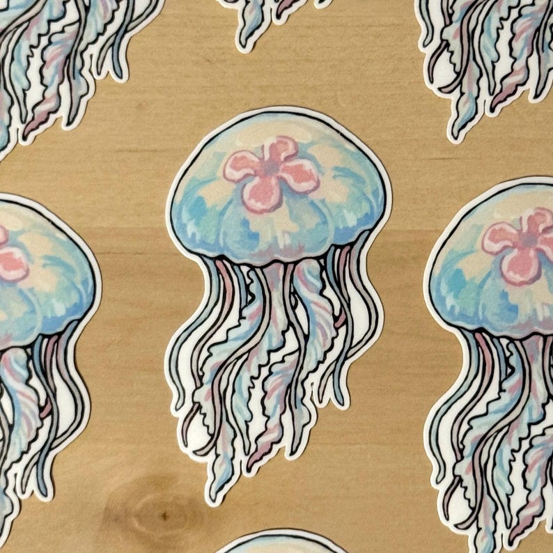 Pu&ograve; includere: Un adesivo fustellato raffigurante una medusa con una campana azzurra traslucida e un centro rosa tenue. La medusa presenta lunghi tentacoli ondulati e fluidi nelle tonalit&agrave; del blu pallido e del rosa, delineati in nero su fondo in legno chiaro.