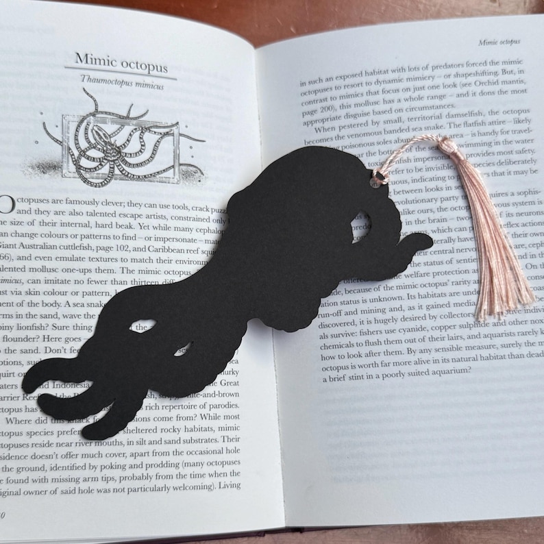 May include: Black octopus-shaped bookmark with a tassel, resting on an open book. The bookmark's design mimics an octopus. The book's pages display text about the mimic octopus.
