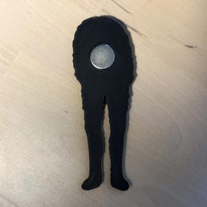 May include: A black, 3D-printed magnet shaped like a pair of long, slender legs extending from a rounded top section. A circular silver-colored metal magnet is embedded in the upper portion of the black textured plastic structure.