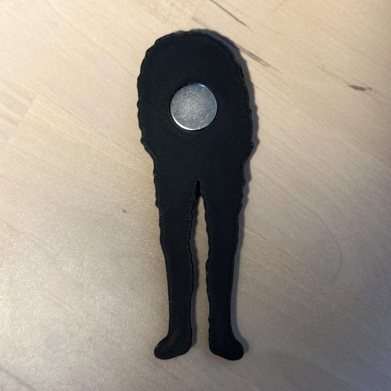 May include: A black, 3D-printed magnet shaped like a pair of long, slender legs extending from a rounded top section. A circular silver-colored metal magnet is embedded in the upper portion of the black textured plastic structure.