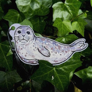 Pode incluir: Um marcador de livros em forma de foca, em estilo aguarela. A foca &eacute; retratada em tons de cinza, branco e roxo, com um contorno preto. Possui uma pequena etiqueta com um borla preta. O fundo &eacute; composto por folhas verdes.