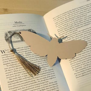 May include: A tan moth-shaped bookmark with a gold tassel rests on an open book. The bookmark is made of thick paper and has a detailed cutout design. The book pages display text about moths.