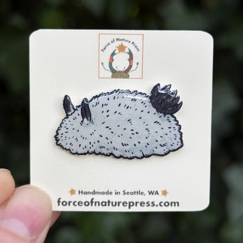 Puede incluir: Un pin de esmalte de nudibranquio gris claro con motas negras y detalles oscuros sobre una tarjeta blanca. La tarjeta incluye el texto Force of Nature Press, un logotipo con un cangrejo, y el texto Handmade in Seattle, WA y forceofnaturepress.com.