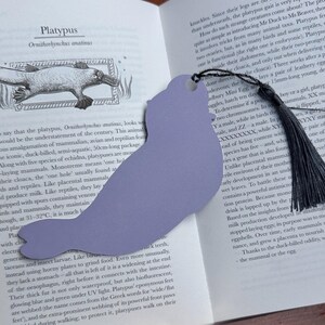 Pode incluir: Um marcador de livros em forma de ornitorrinco, na cor lavanda, com uma borla preta. O marcador de livros est&aacute; colocado num livro com texto sobre ornitorrincos. Um acess&oacute;rio &uacute;nico para os amantes de livros.