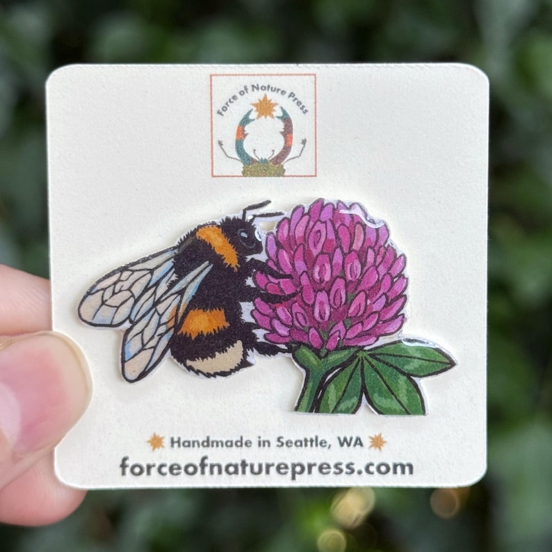 Puede incluir: Un pin que muestra un abejorro peludo con rayas negras y amarillas junto a una flor de tr&eacute;bol rosa vibrante con hojas verdes. El pin est&aacute; sujeto a una tarjeta blanca con el texto Force of Nature Press y Handmade in Seattle, WA.