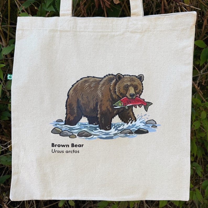 May include: A cream-colored canvas tote bag featuring a graphic illustration of a brown bear standing in a stream and holding a red salmon in its mouth. Below the illustration, the text reads Brown Bear and the scientific name Ursus arctos.