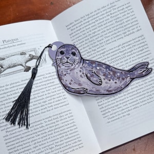Pode incluir: Um marcador em forma de foca com um borla preta, pousado num livro aberto. O marcador &eacute; roxo claro com uma ilustra&ccedil;&atilde;o detalhada de uma foca. As p&aacute;ginas do livro exibem texto e ilustra&ccedil;&otilde;es.