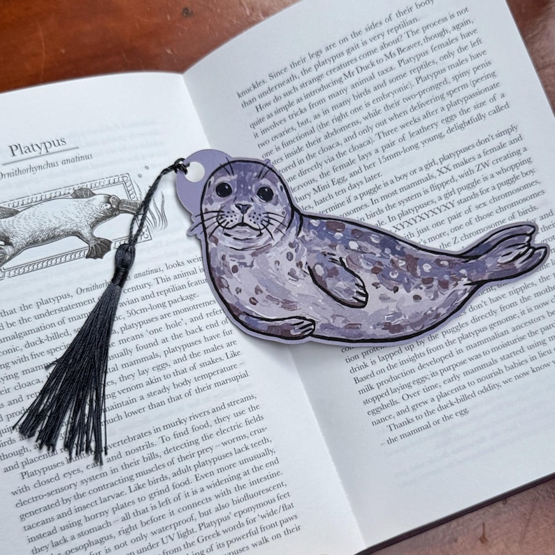 Pode incluir: Um marcador em forma de foca com um borla preta, pousado num livro aberto. O marcador &eacute; roxo claro com uma ilustra&ccedil;&atilde;o detalhada de uma foca. As p&aacute;ginas do livro exibem texto e ilustra&ccedil;&otilde;es.