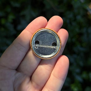 May include: A circular pin-back button featuring a metallic silver backing with a horizontal safety pin closure mechanism. The outer edge of the button is orange, and the central circular area has a textured, dark gray, speckled surface finish.