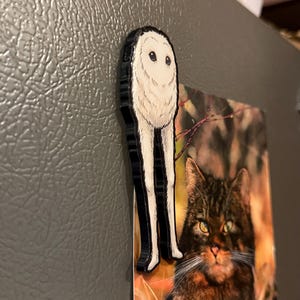 May include: A flat, die-cut magnet featuring a stylized, fluffy white creature with a round head, large dark eyes, and two long, slender legs. The magnet is attached to a gray metallic surface, partially overlapping a rectangular photograph of a cat.