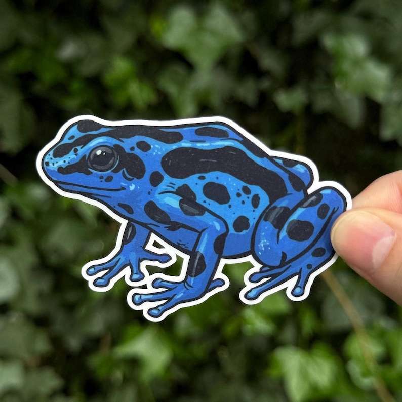 May include: A die-cut sticker featuring a vibrant blue poison dart frog with distinct black spots and patches across its body. The illustration shows the frog in a side profile view with a white border outline, showcasing its smooth skin texture.