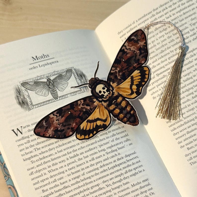 May include: A decorative bookmark featuring a detailed illustration of a Death's Head Hawkmoth with a skull design on its thorax. The bookmark is set against the pages of a book with the title "Moths" visible.