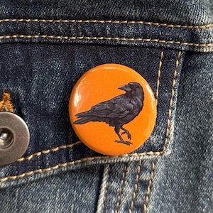 May include: A round, orange button featuring a detailed black illustration of a raven. The button is pinned to a blue denim jacket, with visible stitching and a metal button clasp.