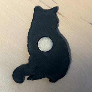 Puede incluir: Un im&aacute;n negro con forma de gato y superficie texturizada. La silueta muestra un gato sentado de perfil, mirando hacia la derecha, con un disco magn&eacute;tico met&aacute;lico de color plateado incrustado en el centro del cuerpo como base magn&eacute;tica funcional.