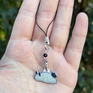 Puede incluir: Un peque&ntilde;o dije de babosa de mar con un dise&ntilde;o en blanco y negro, sujeto a un cord&oacute;n negro con un cierre plateado y una cuenta negra. El dije est&aacute; sostenido por una mano.