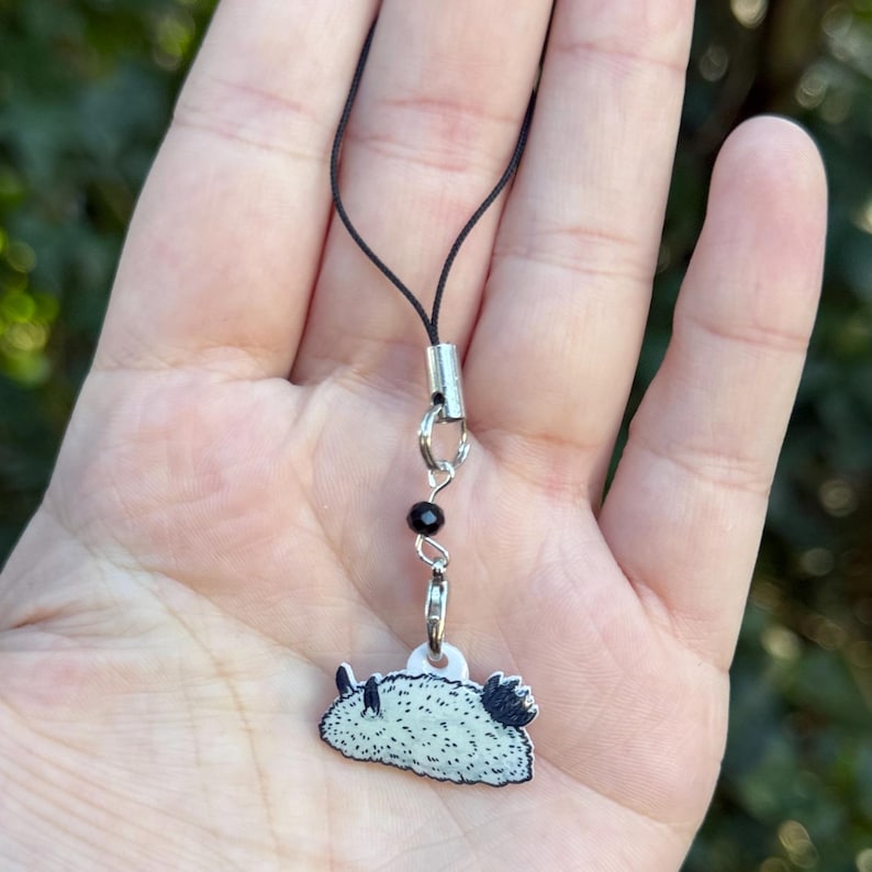 Puede incluir: Un peque&ntilde;o dije de babosa de mar con un dise&ntilde;o en blanco y negro, sujeto a un cord&oacute;n negro con un cierre plateado y una cuenta negra. El dije est&aacute; sostenido por una mano.