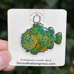 Puede incluir: Un pin de esmalte brillante con forma de pez redondo y espinoso con manchas verdes y amarillas. El pin est&aacute; sujeto a una tarjeta blanca que presenta el texto Force of Nature Press, un logotipo y la direcci&oacute;n web forceofnaturepress.com.
