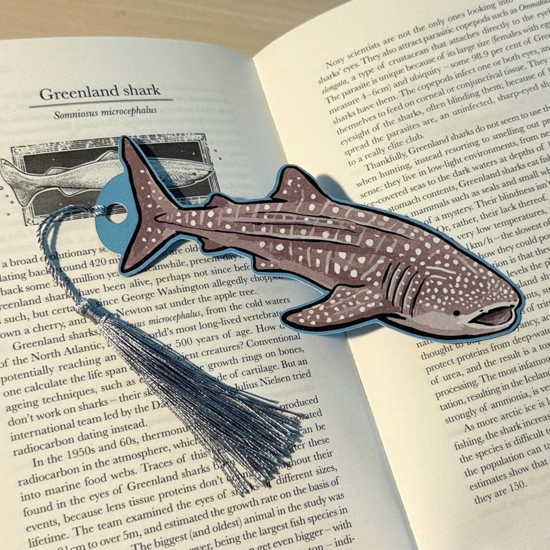 May include: A whimsical bookmark featuring a detailed illustration of a Greenland shark, showcasing its distinctive spotted pattern and shape. The bookmark has a blue accent and a silver tassel, perfect for marking pages in a book.