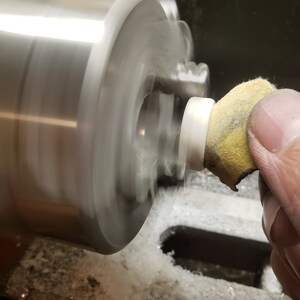 May include: A close-up shot of a lathe in operation, with a white cylindrical object being worked on. A hand holds a yellow and tan abrasive pad against the spinning piece. The machine is silver and the background is dark.