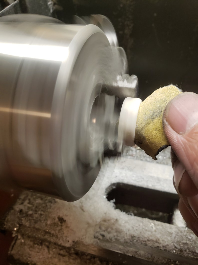 May include: A close-up shot of a lathe in operation, with a white cylindrical object being worked on. A hand holds a yellow and tan abrasive pad against the spinning piece. The machine is silver and the background is dark.