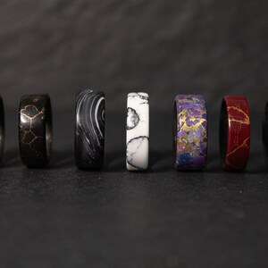 May include: Eight different colored rings with unique patterns. The rings are made of a dark material with gold accents. The rings are arranged in a row from left to right.