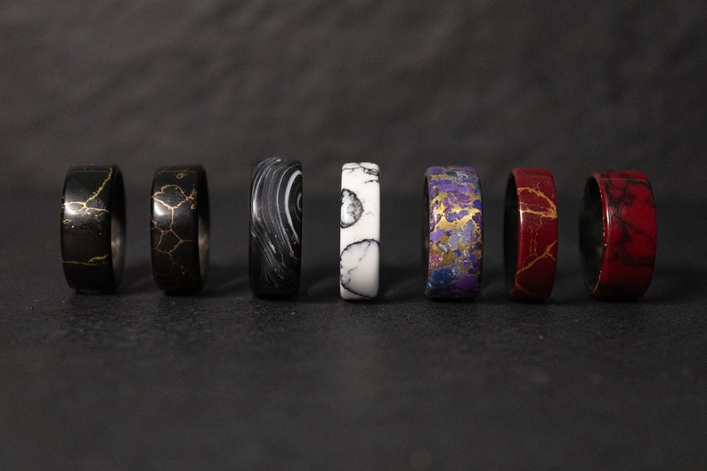 May include: Eight different colored rings with unique patterns. The rings are made of a dark material with gold accents. The rings are arranged in a row from left to right.
