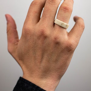 May include: A rectangular ring made of a light-colored material, possibly stone or bone, is worn on a finger. The ring has a smooth, flat surface and a simple, minimalist design. The hand is in focus, with a black jacket sleeve in the background.