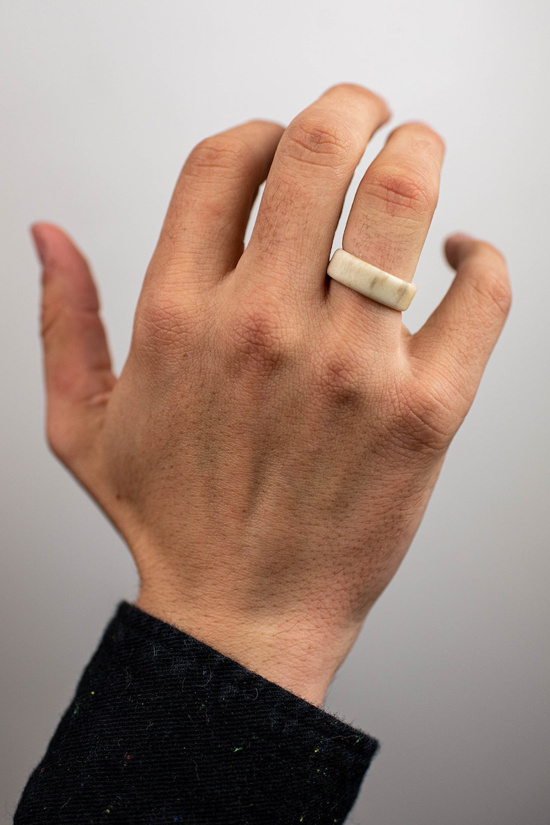 May include: A rectangular ring made of a light-colored material, possibly stone or bone, is worn on a finger. The ring has a smooth, flat surface and a simple, minimalist design. The hand is in focus, with a black jacket sleeve in the background.
