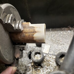 May include: A close-up of a lathe machine in operation, with a piece of wood being shaped. The wood is held in a chuck, and a cutting tool is visible, removing material. The wood has a light brown and cream color.