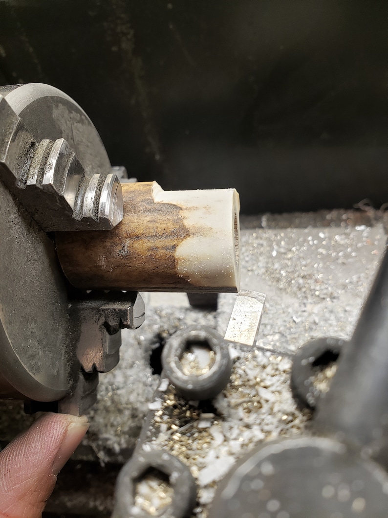 May include: A close-up of a lathe machine in operation, with a piece of wood being shaped. The wood is held in a chuck, and a cutting tool is visible, removing material. The wood has a light brown and cream color.