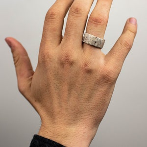 May include: A wide, rectangular ring made of a light-colored material with a speckled pattern. The ring is worn on a finger, showcasing its unique texture and design. The ring's width is approximately 0.75 inches.