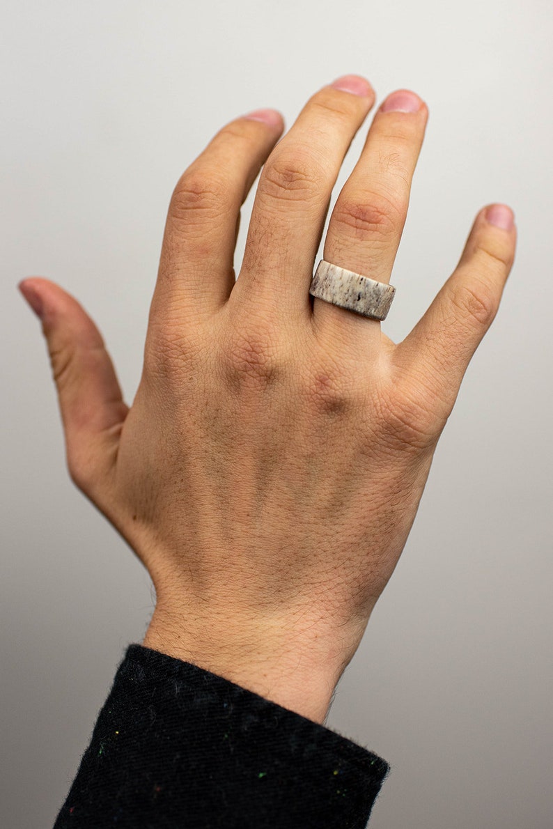 May include: A wide, rectangular ring made of a light-colored material with a speckled pattern. The ring is worn on a finger, showcasing its unique texture and design. The ring's width is approximately 0.75 inches.