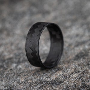 Op de afbeelding: Een zwarte carbonfiber ring met een geweven patroon. De ring is glad en gepolijst.