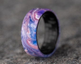May include: A close-up of a ring with a marbled design. The ring features a swirling pattern of purple, blue, and pink hues on the outer band. The inner band is black. The ring is set against a gray, textured background.