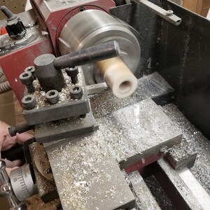May include: A close-up of a metal lathe in operation, with a cylindrical piece of light-colored material being shaped. The machine is red and silver, with metal shavings visible around the work area. The lathe is actively spinning the material.