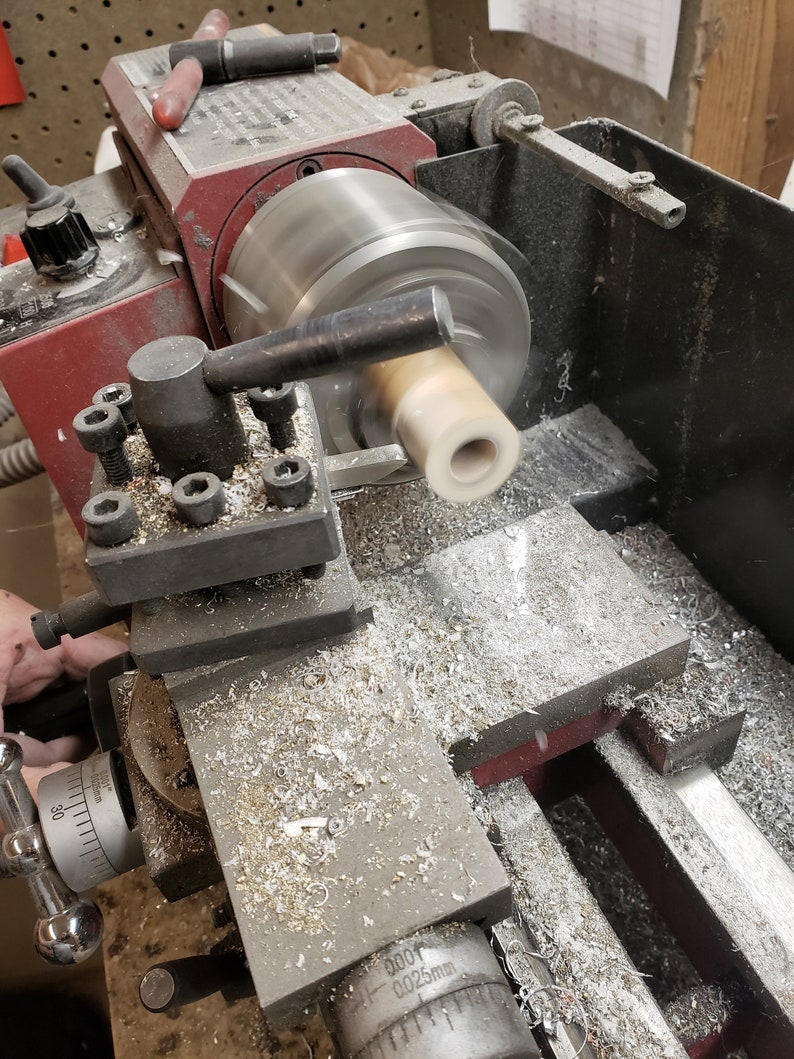 May include: A close-up of a metal lathe in operation, with a cylindrical piece of light-colored material being shaped. The machine is red and silver, with metal shavings visible around the work area. The lathe is actively spinning the material.