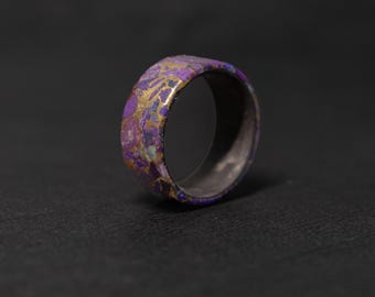May include: A close-up of a wide band ring with a unique design. The ring features a vibrant mix of purple, gold, and teal colors. The interior of the ring is a dark gray. The ring is set against a dark background.