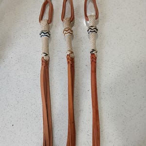 May include: Three tan leather whips featuring a braided handle section with a decorative knot pattern in black and white. Each whip has a looped top and a series of long, thin leather strips extending from the base of the braided handle.