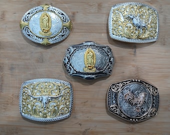 May include: Five silver and gold-toned metal belt buckles with various designs. The buckles feature religious figures, a bull, and a rooster. The buckles are displayed on a wooden surface.
