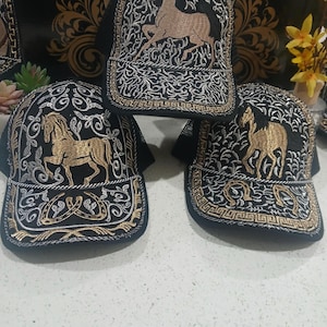 May include: Three black trucker hats with embroidered horse designs. The hats feature gold and silver thread detailing, including a running horse on the front panel and ornate patterns. The brims are black, and the hats are displayed on a white surface.