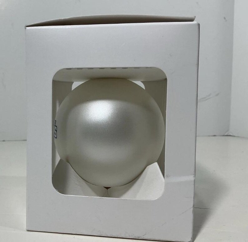 May include: A frosted, spherical ornament is displayed inside a white cardboard box. The ornament has a subtle sheen and a small, dark logo on its surface. The box has a square opening that frames the ornament.