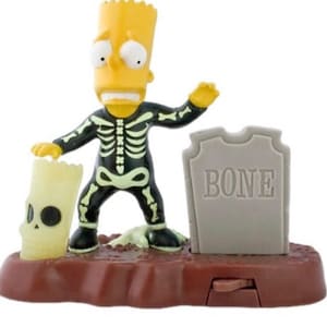 May include: A Bart Simpson figurine in a skeleton costume stands on a brown base with a tombstone that reads "BONE." The figurine holds a skull-shaped object. The figure is yellow, black, and white.