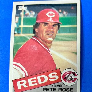 May include: A baseball card featuring Pete Rose in a red Cincinnati Reds uniform and cap. The card has the word "REDS" in large white letters on a red background. The card also includes the team logo and the text "1B-MGR PETE ROSE".