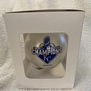 May include: A baseball in a white box. The baseball is white with the text "2015 WORLD SERIES CHAMPIONS KC" in blue and white. The box is white with cut-out sections.