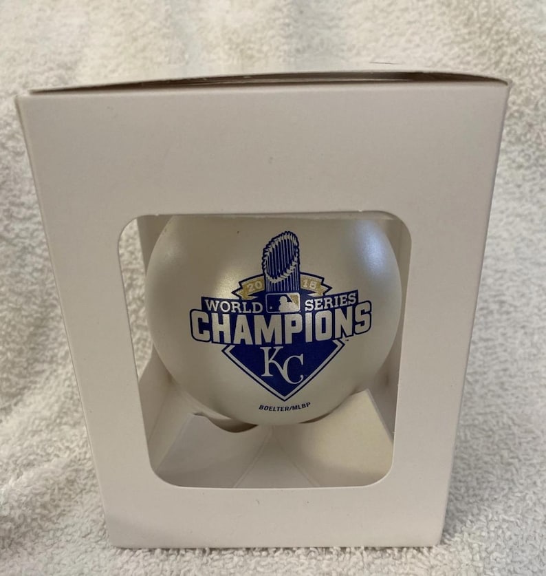 May include: A baseball in a white box. The baseball is white with the text "2015 WORLD SERIES CHAMPIONS KC" in blue and white. The box is white with cut-out sections.
