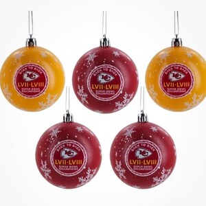 May include: Five Christmas ornaments. Two are yellow, and three are red. Each ornament has the text "BACK TO BACK LVII-LVIII SUPER BOWL CHAMPIONS" in a circular design. White snowflakes are scattered across the ornaments.